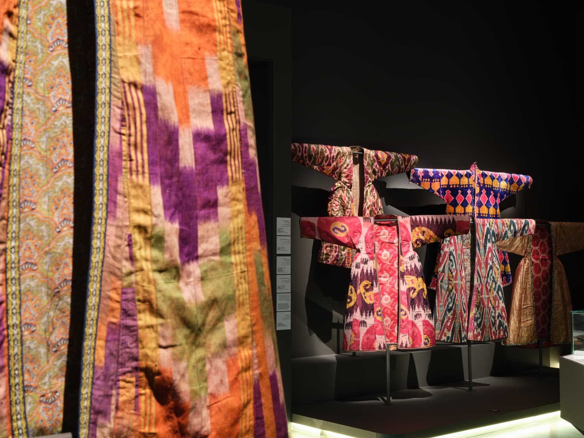 Brightly patterned Uzbek ikat textiles and garments displayed in a dark gallery